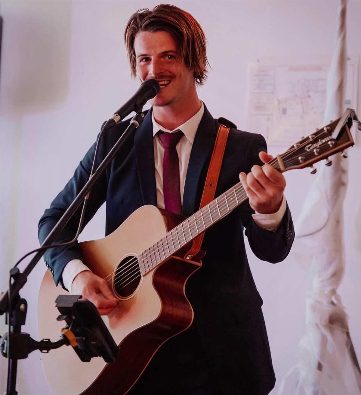Jake Wood smiling as he sings and plays acoustic guitar in a suit